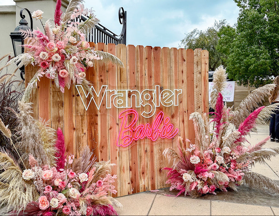 Decorative setup with flowers and 'Wrangler Barbie' sign in front of a wooden fence.